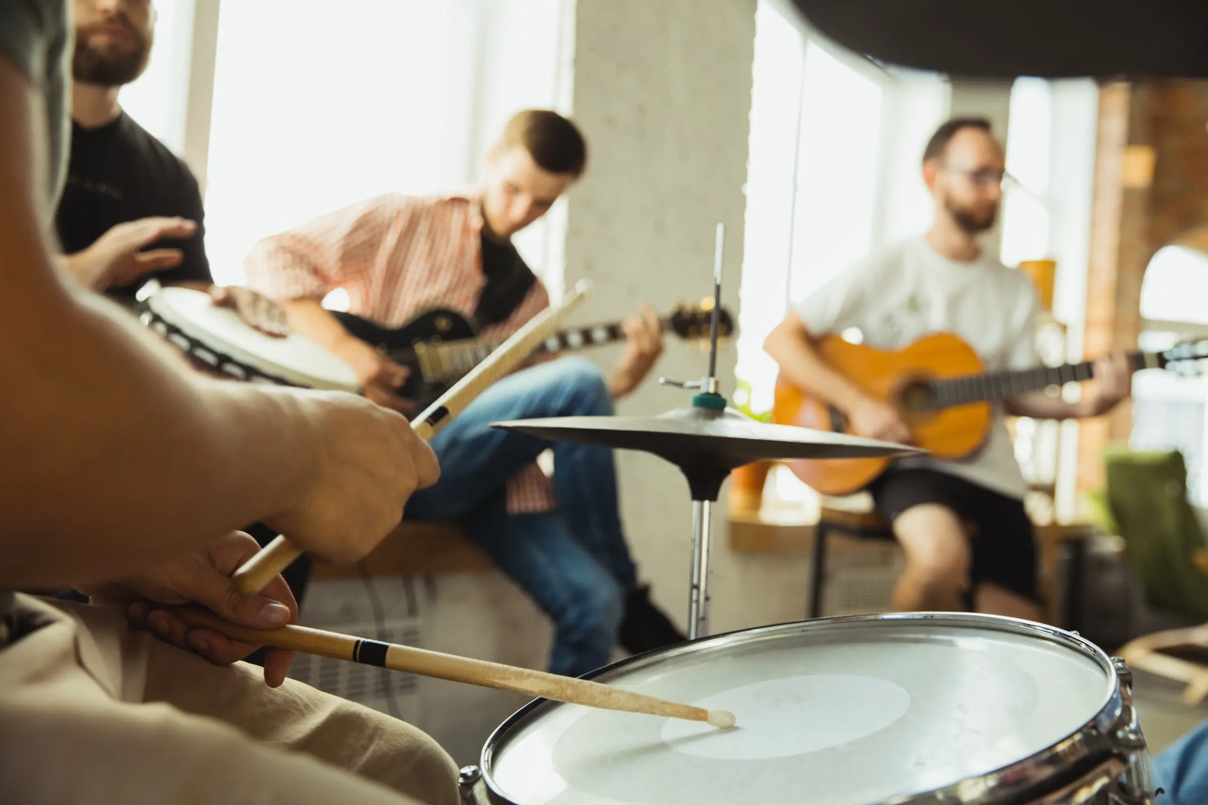 Musicians rehearsing together with drums and guitars in a bright room.