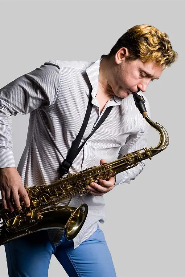 Blond man playing tenor saxophone against a grey background