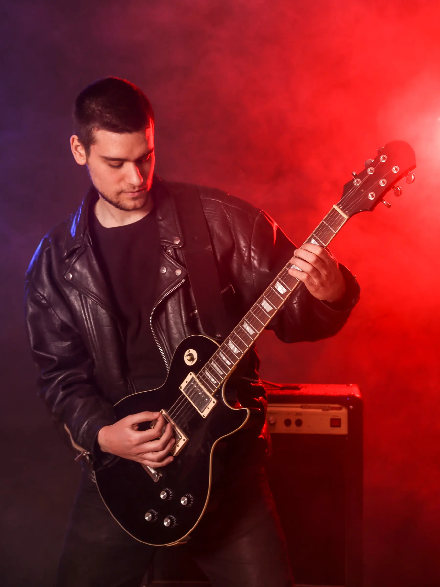 Young guitarist in a leather jacket playing an electric guitar in red light.