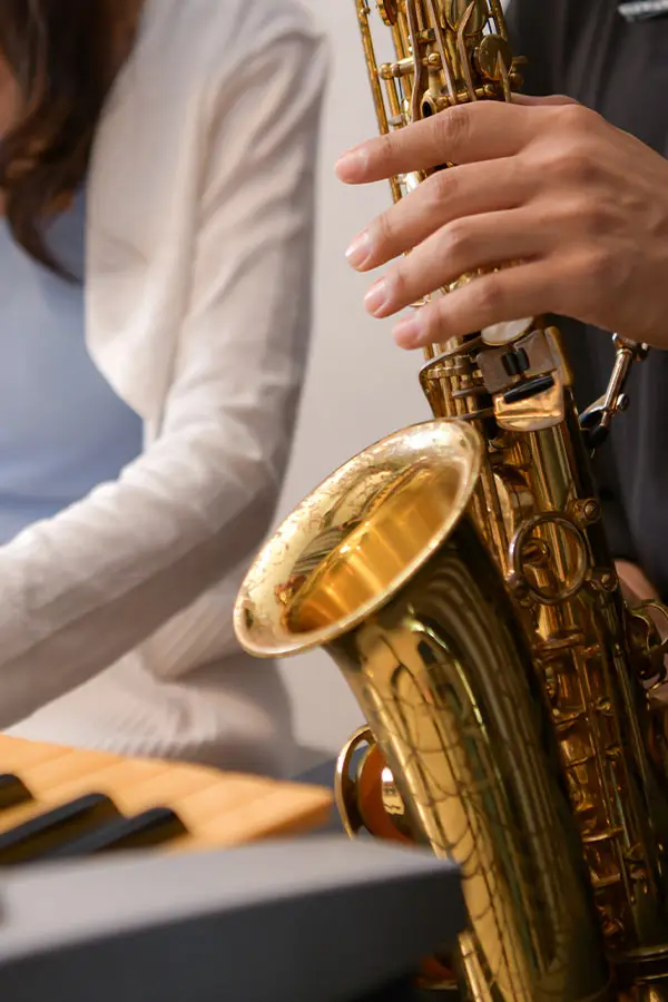 Hands holding a saxophone beside a seated person in a white cardigan
