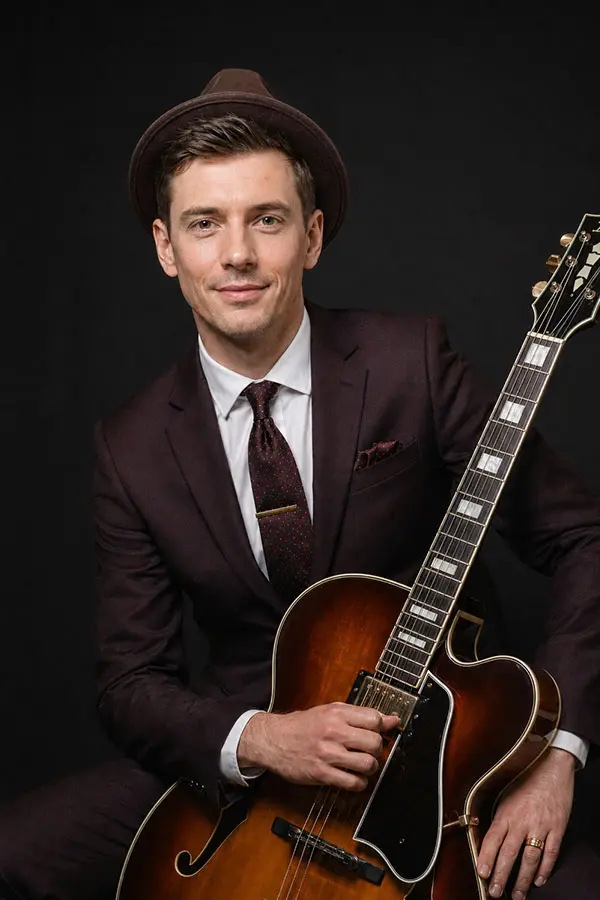 Man in a hat and suit holding an archtop electric guitar
