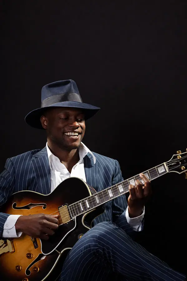 Smiling man in a fedora playing a hollow-body electric guitar
