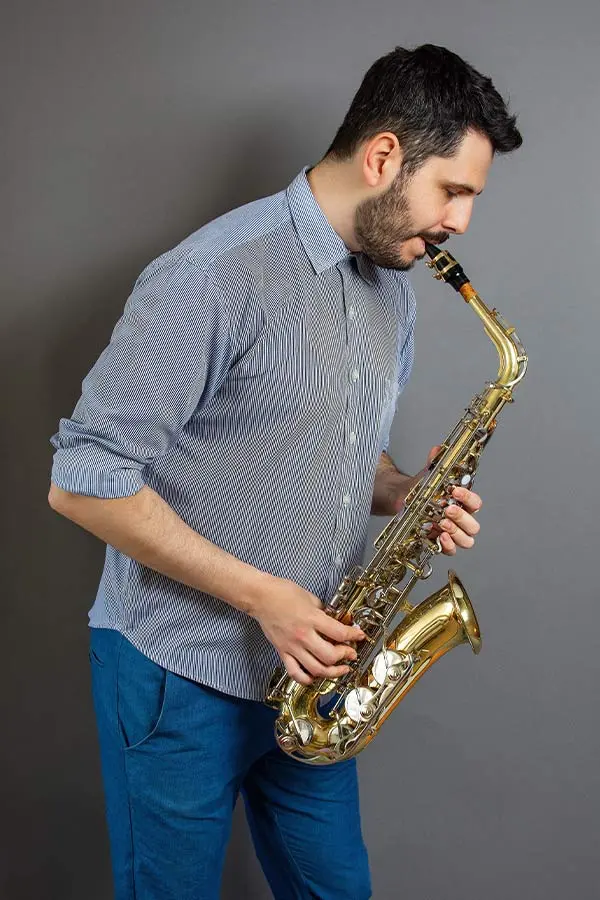 Man in blue trousers playing alto saxophone against a grey background