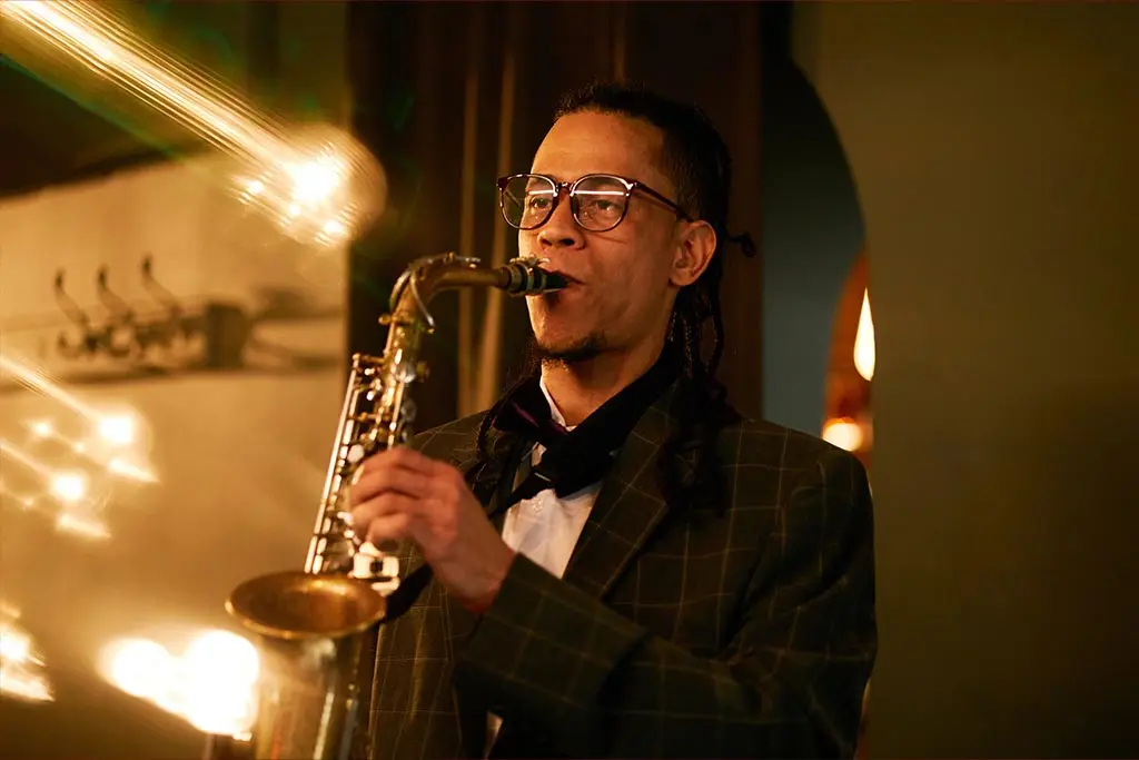 Man with glasses playing alto saxophone under warm string lights