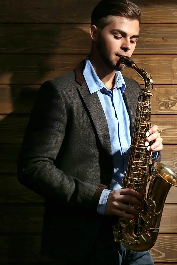 Man in a blazer playing alto saxophone by a wooden wall