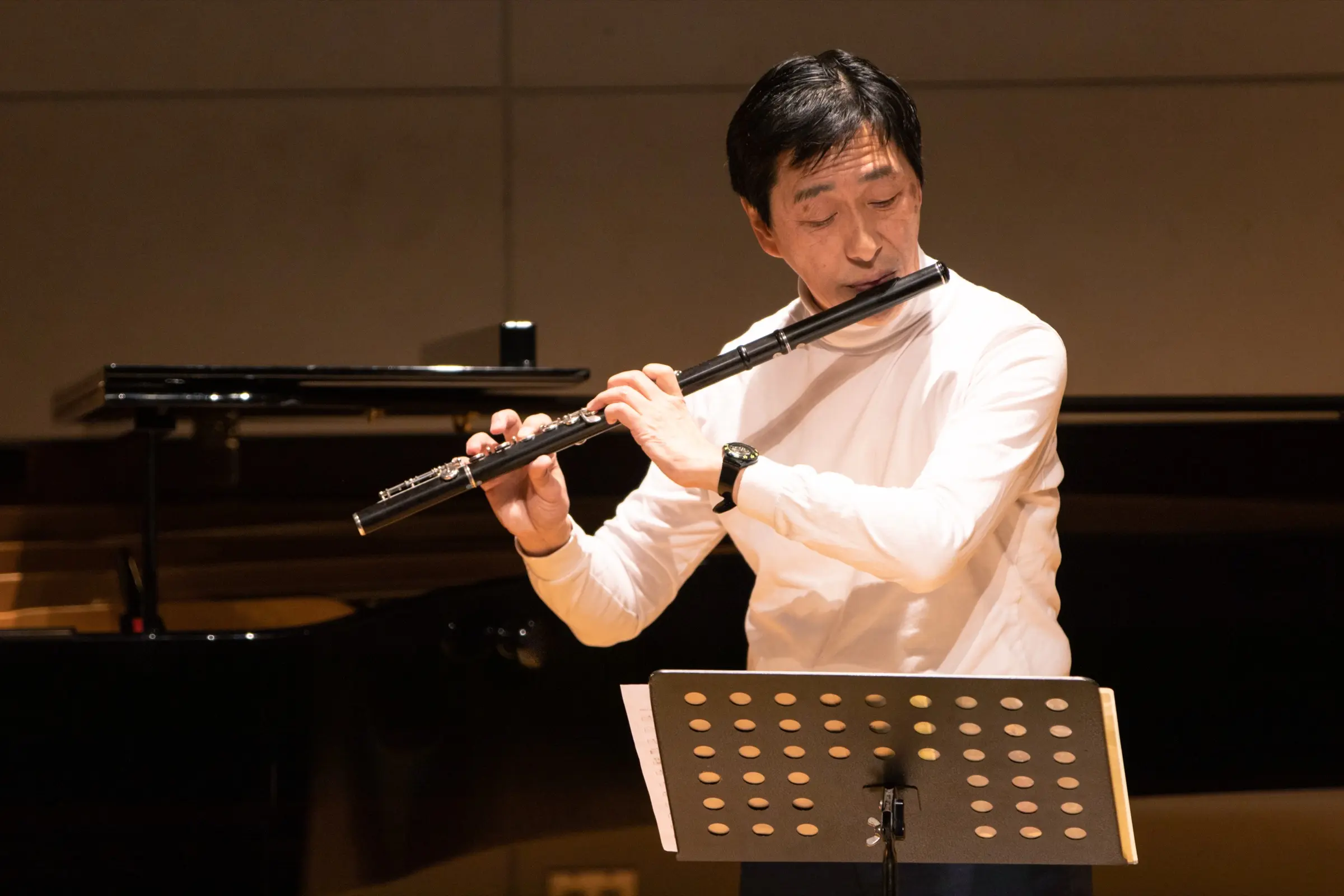 A man playing the flute at a music stand beside a grand piano