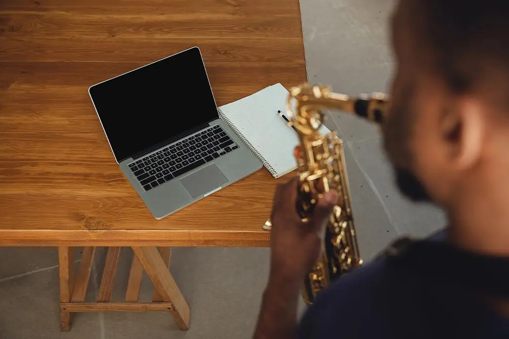 Man playing saxophone beside a laptop and notebook on a wooden table