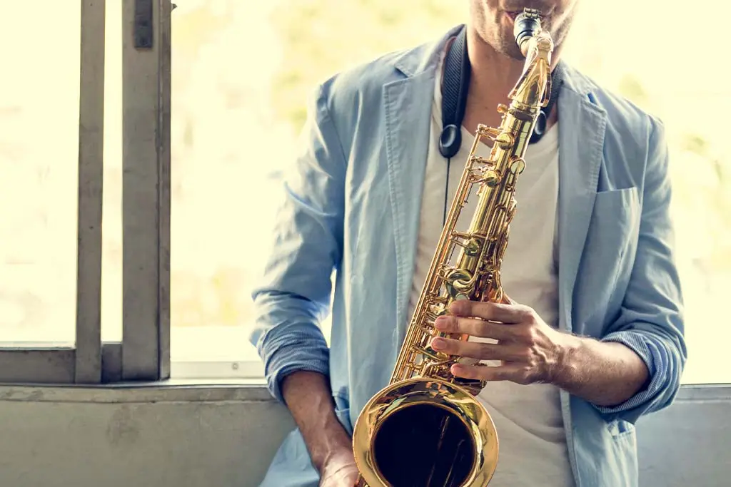 Man in a blue jacket playing saxophone beside a window