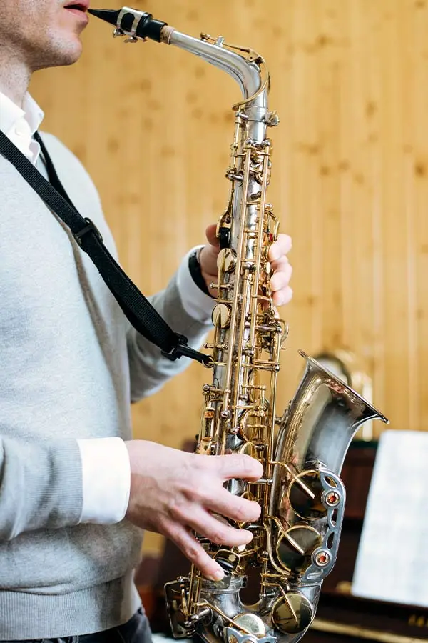 Man in a grey jumper playing saxophone by a wooden wall