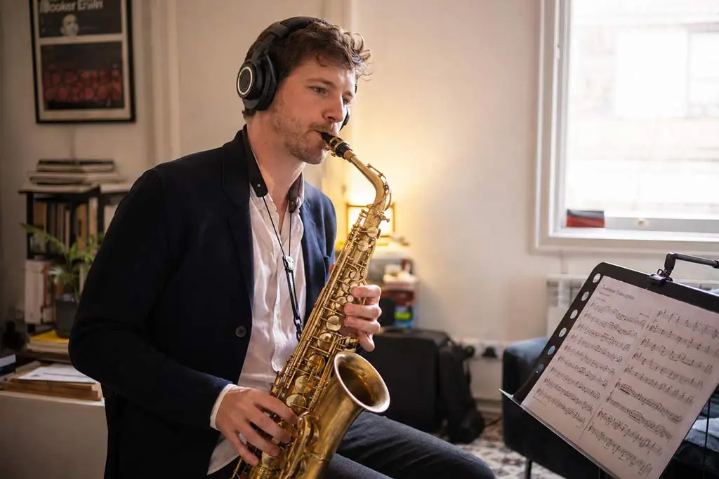 Man wearing headphones playing tenor saxophone beside sheet music