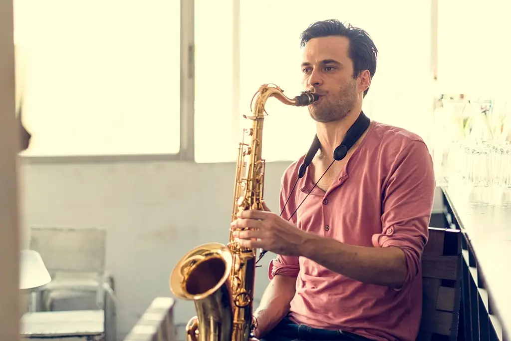 Man in a pink shirt playing tenor saxophone beside a window