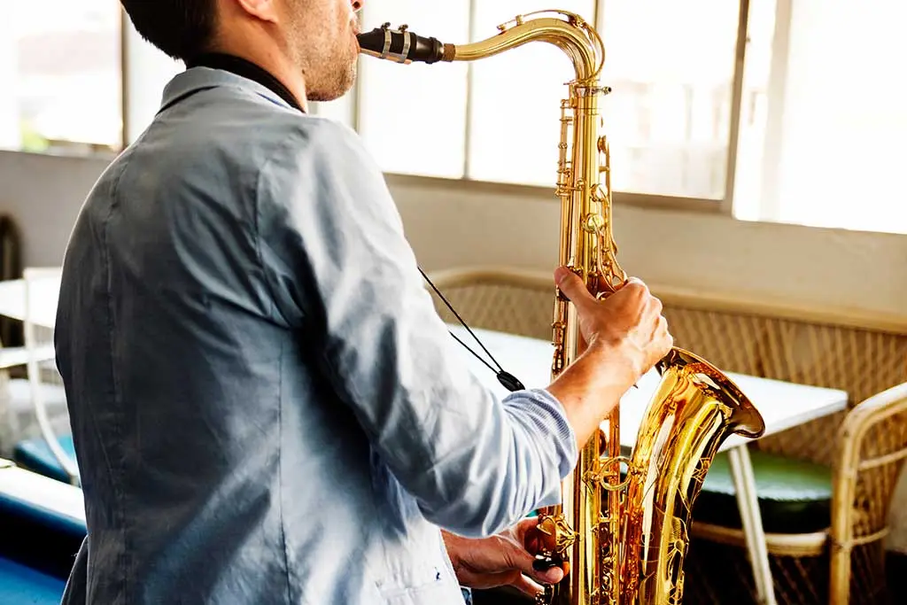 Man in a blue shirt playing tenor saxophone indoors by windows