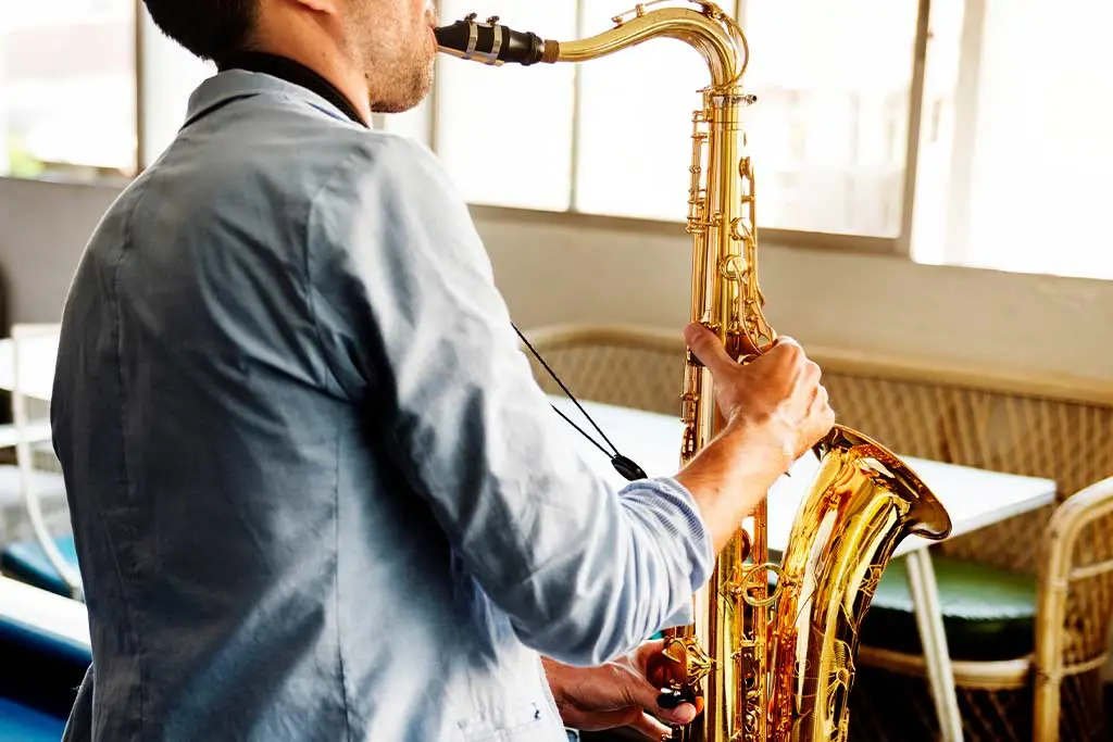 Man in a blue shirt playing tenor saxophone by windows