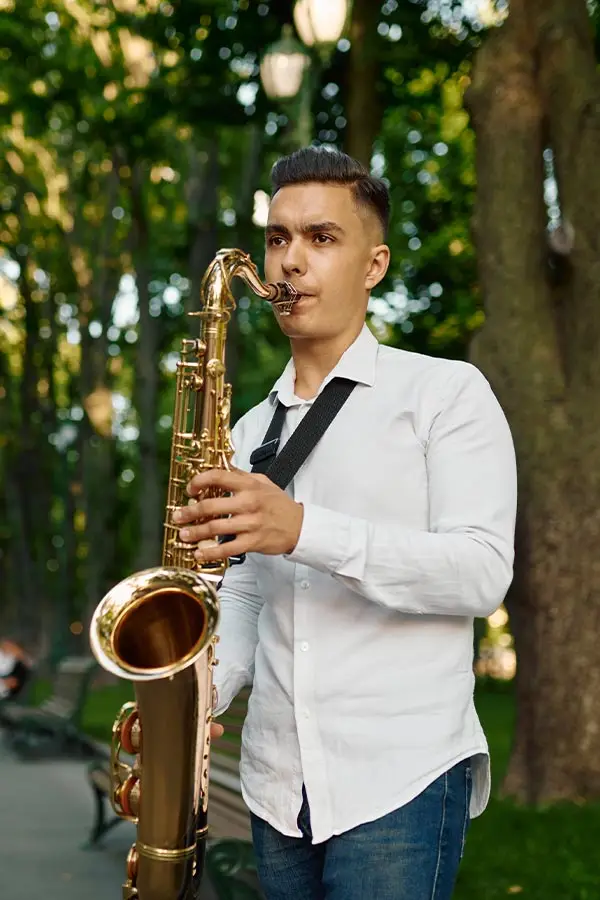 Man in a white shirt playing tenor saxophone under trees