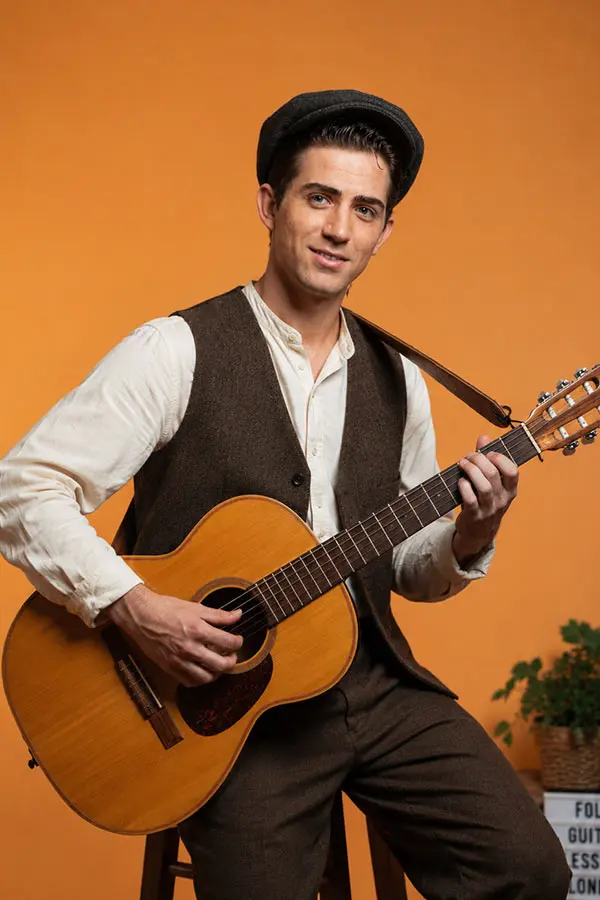 Man seated with an acoustic guitar against an orange background