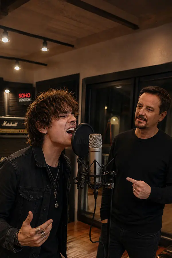Man singing into a studio microphone while another man gestures beside him