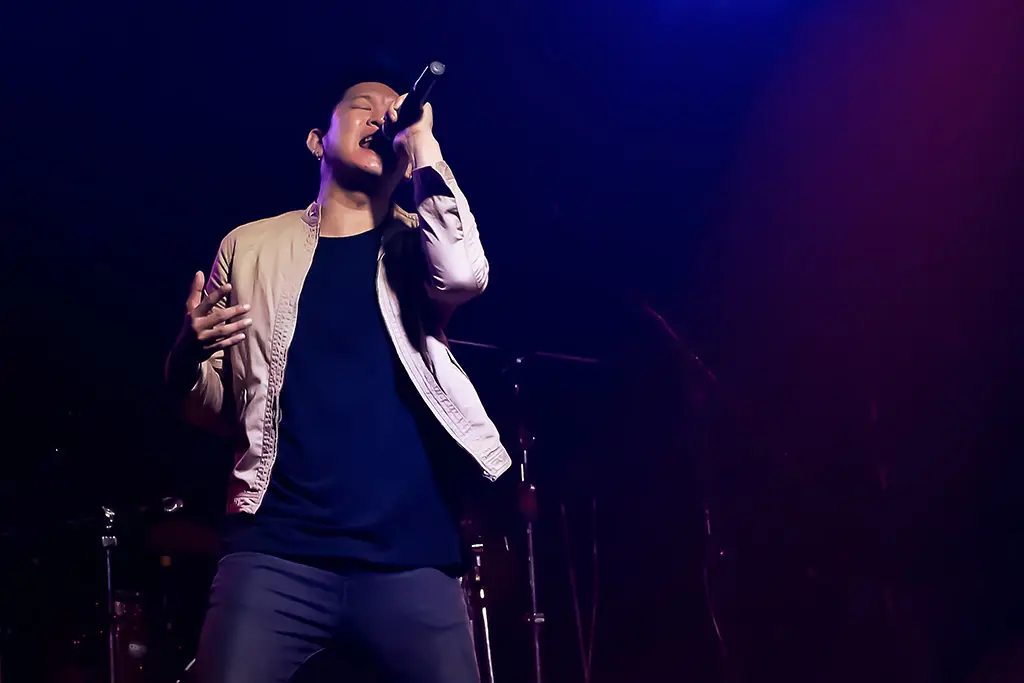 Man singing into a handheld microphone under purple stage lights