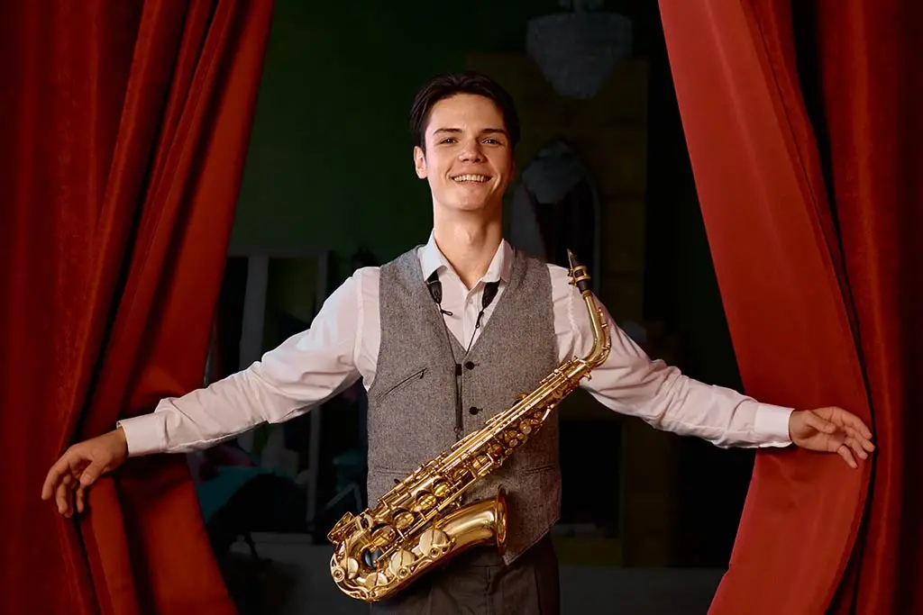 Man holding an alto saxophone between red curtains