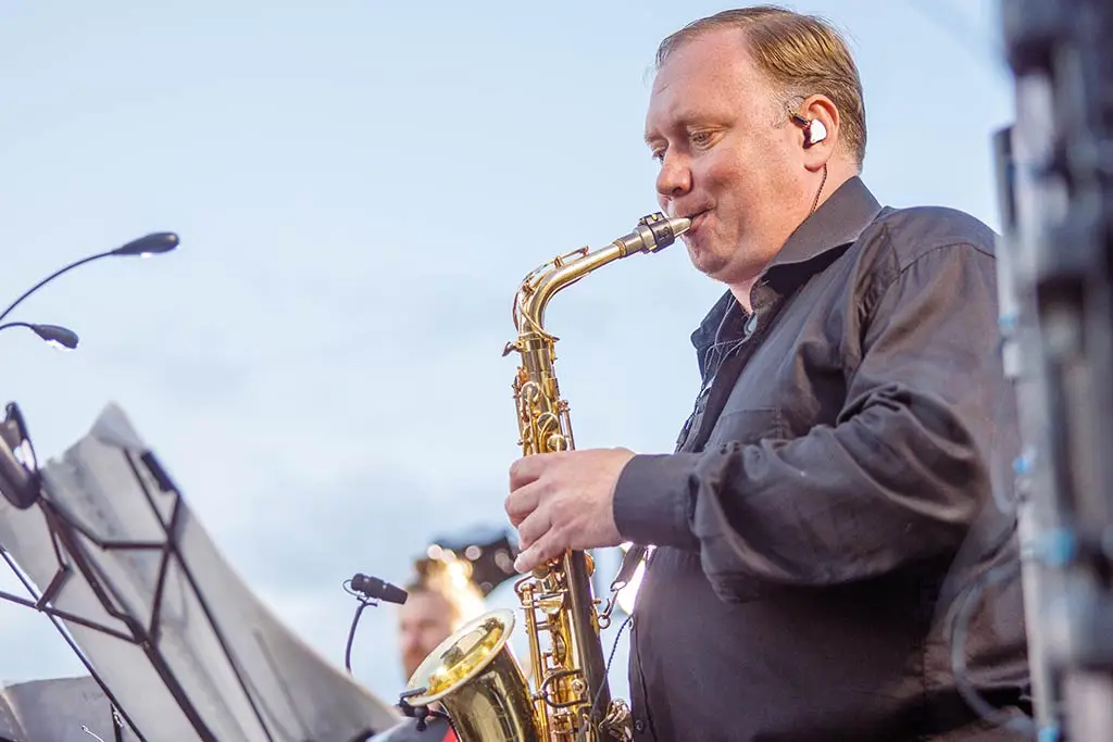 Older man in a dark shirt playing saxophone beside a music stand