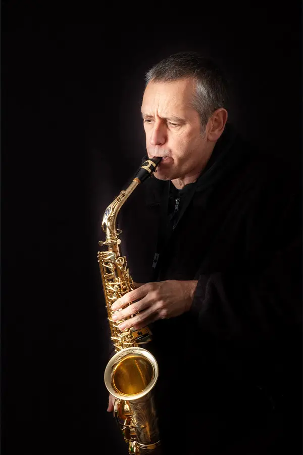Older man playing alto saxophone against a black background
