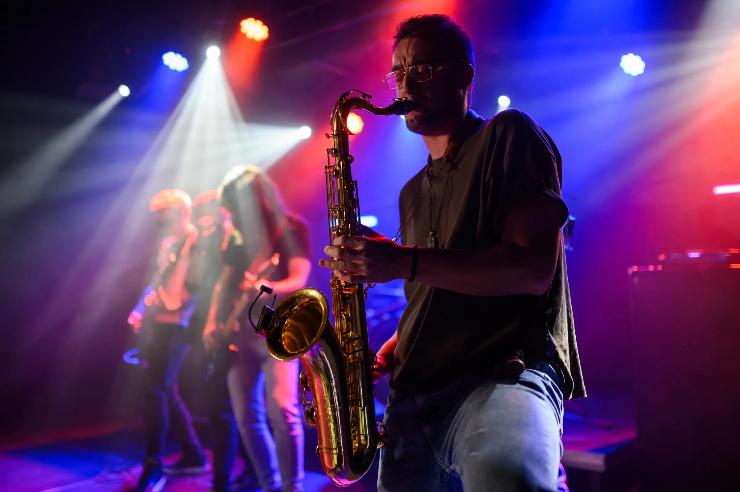 Saxophonist playing with a band under colourful stage lights.