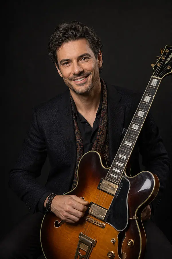 Smiling man in a black blazer holding a sunburst electric guitar