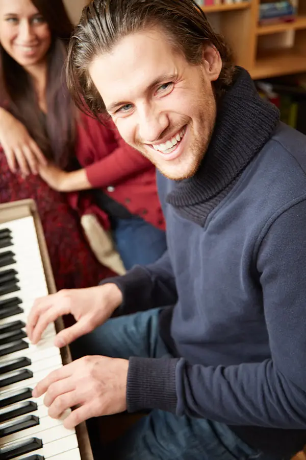 Smiling man in a blue jumper playing piano as a woman sits beside him