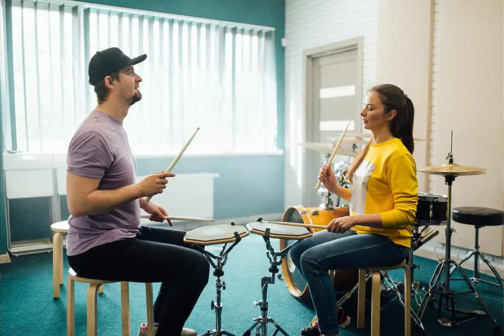 Two adults holding drumsticks beside practice pads and a drum kit
