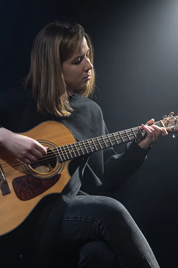 Woman seated and playing an acoustic guitar against a dark background