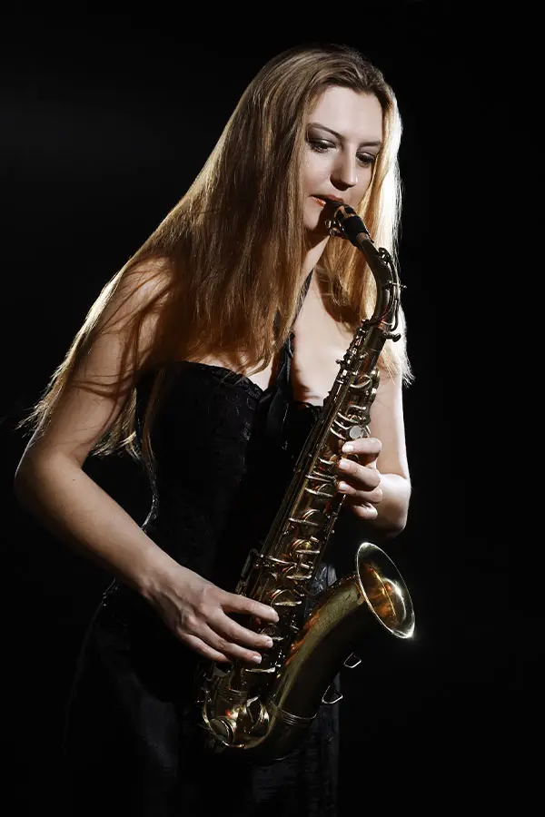 Woman in a black dress playing an alto saxophone against darkness
