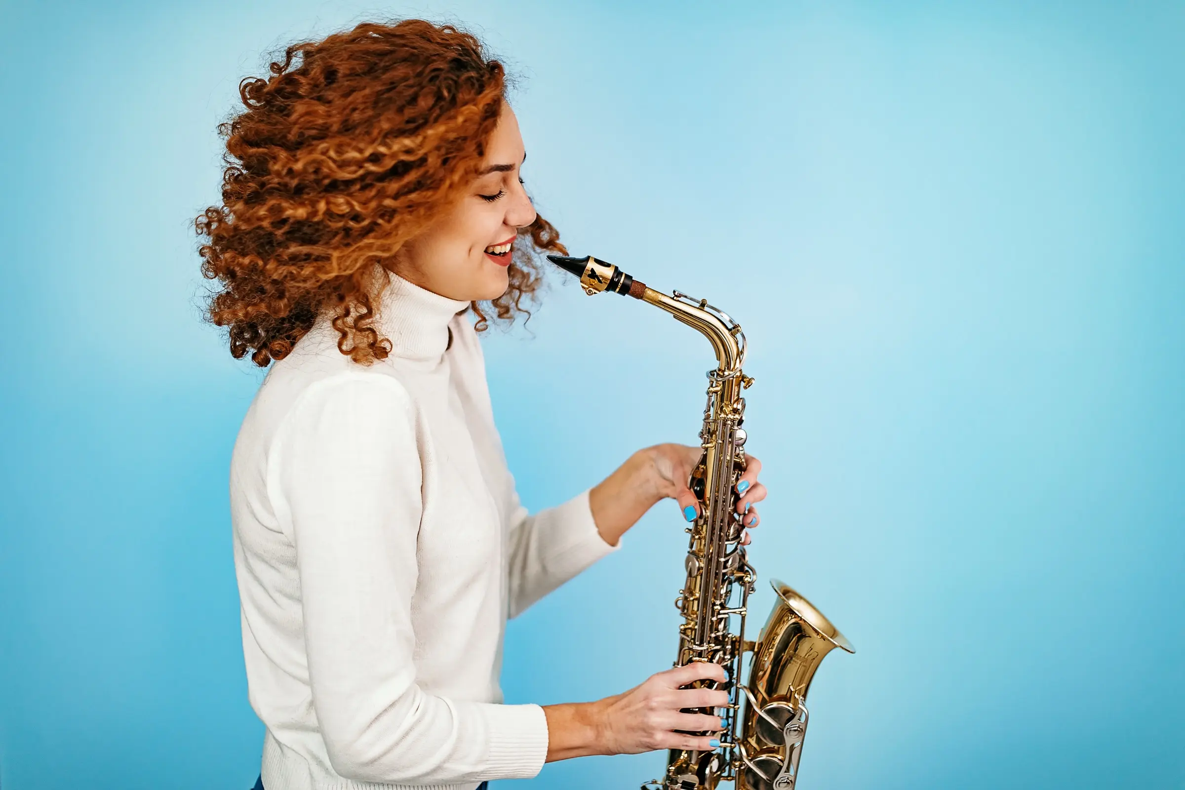 Woman with curly red hair playing an alto saxophone against a blue background.