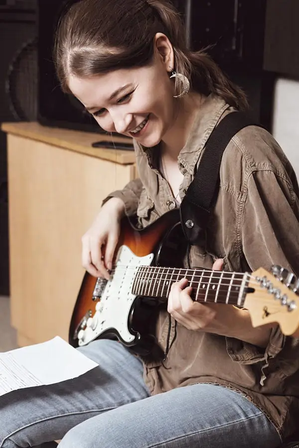 Smiling woman playing an electric guitar with sheet music nearby