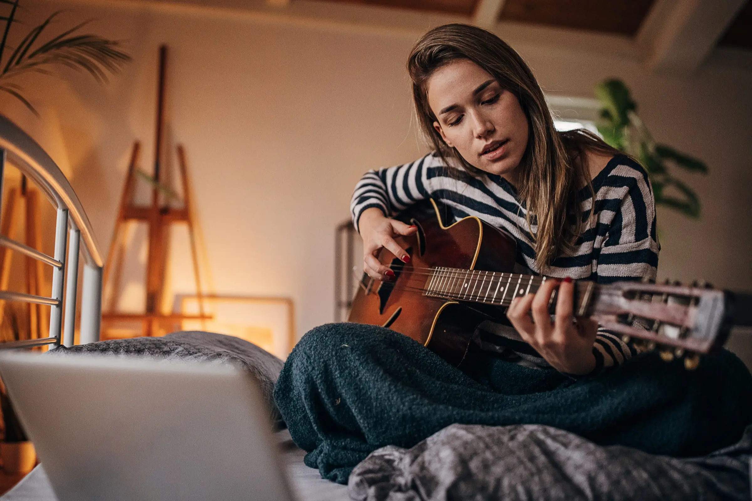 Woman in a striped top playing an acoustic guitar on her bed.