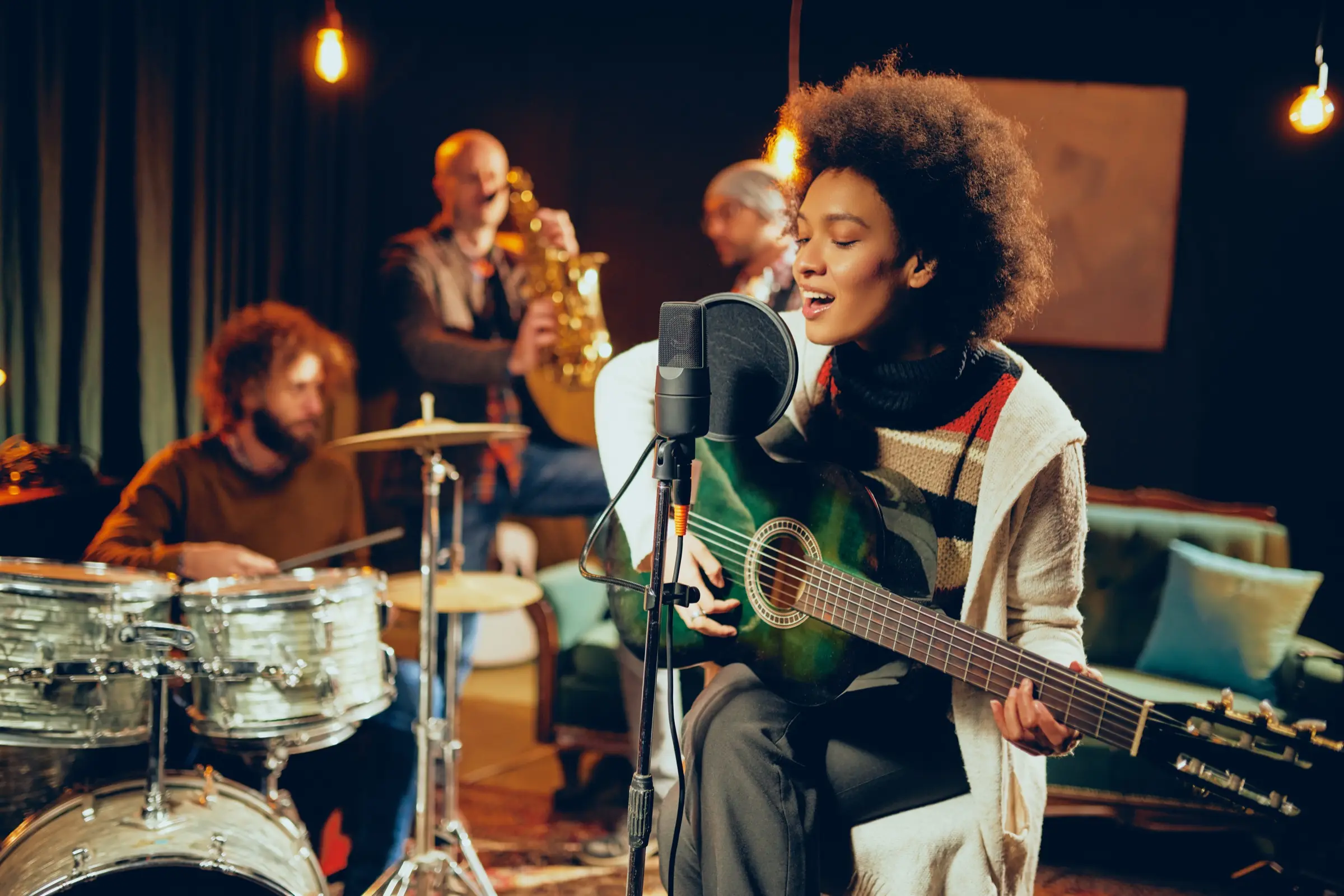 A woman singing and playing guitar with drums and saxophone behind her