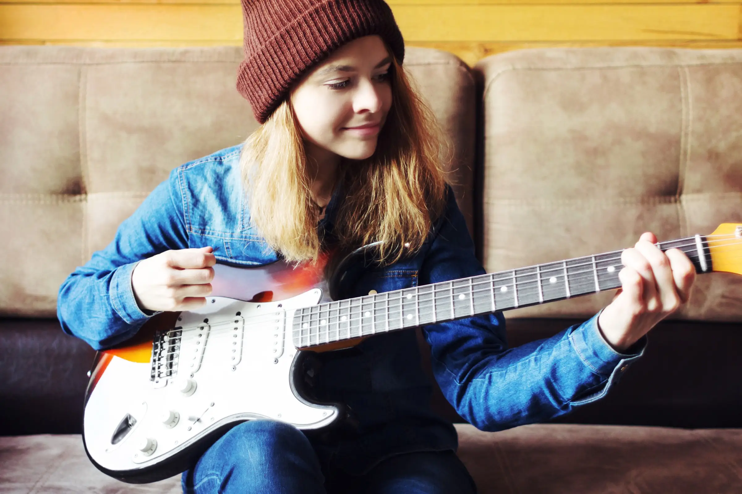 A young woman in a beanie playing an electric guitar on a sofa