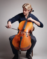 A blond man playing the cello, seated in a studio
